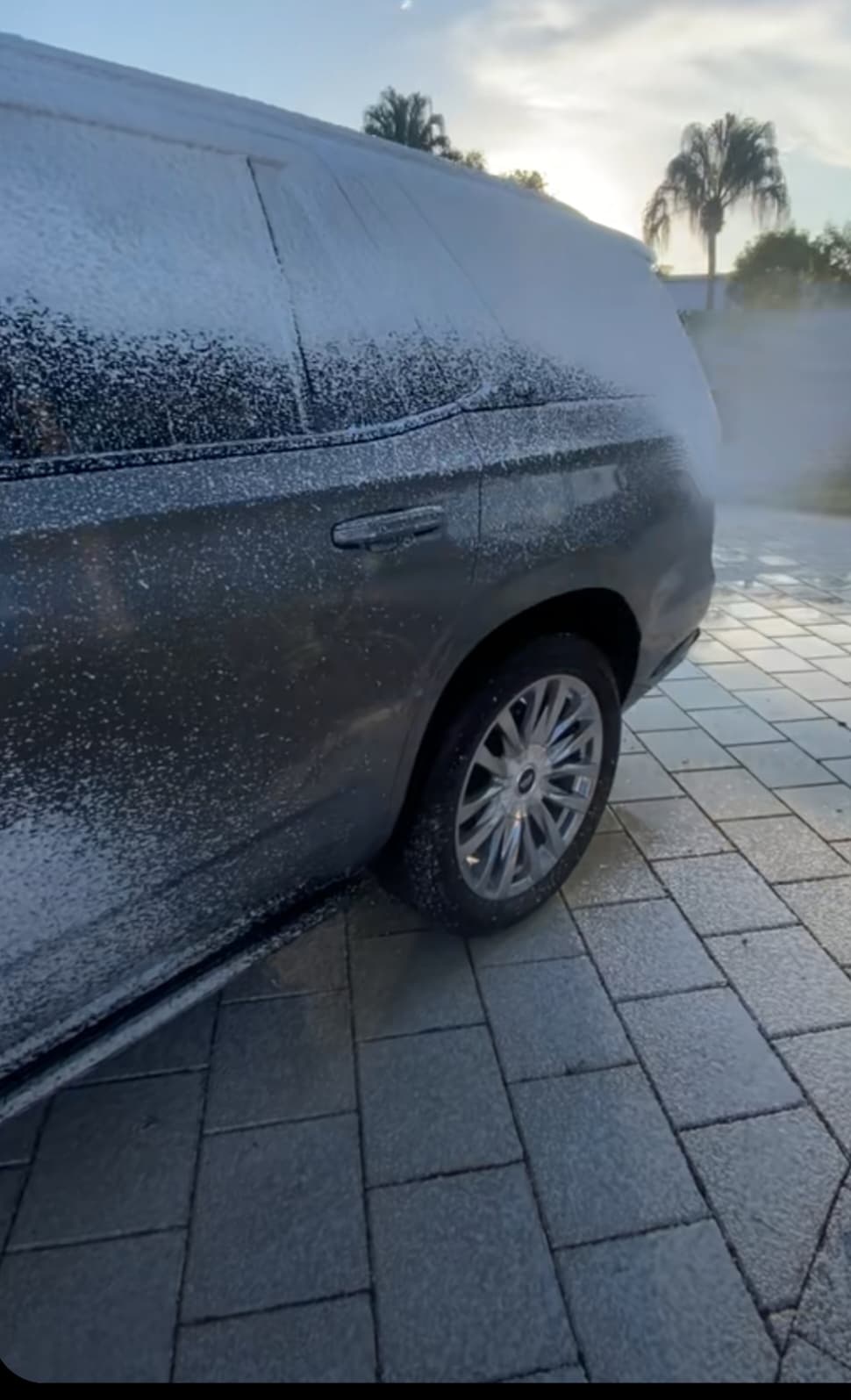 Thick white car wash foam covers a gray SUV parked on a paved stone driveway.