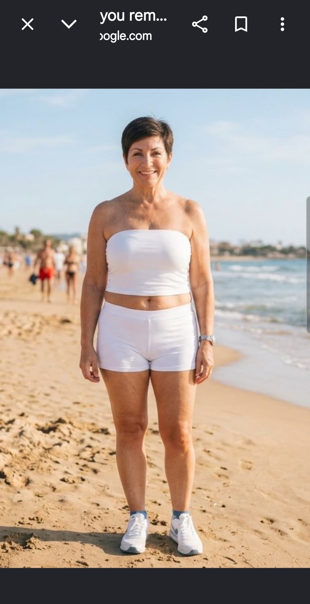 Smiling middle-aged woman in white athletic wear standing on a sunny, sandy beach.