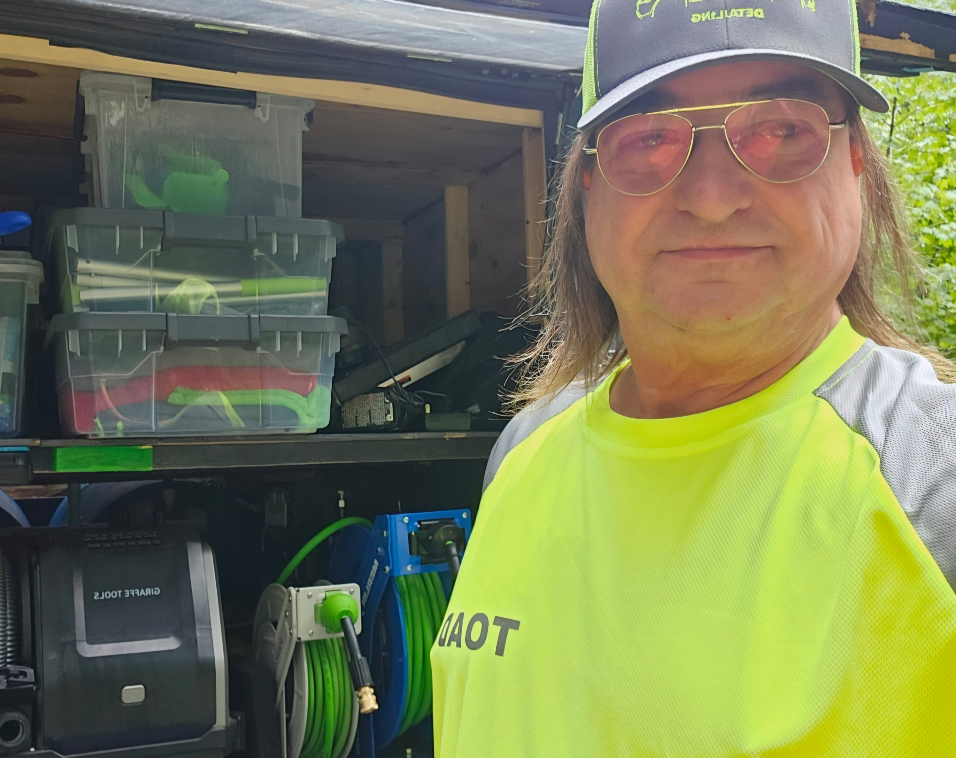 Man in neon shirt and cap stands before a truck filled with detailing equipment.