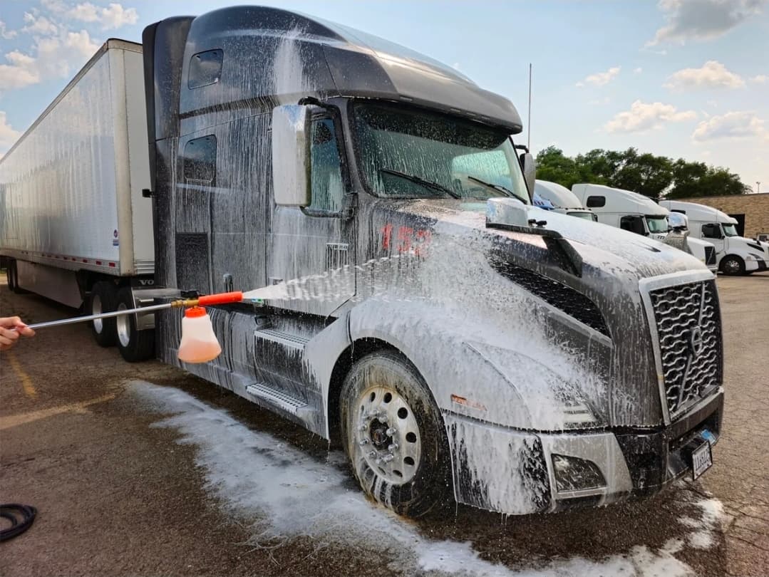 Thick white cleaning foam being sprayed onto a dark grey Volvo semi-truck from a handheld nozzle.