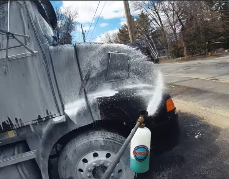 Thick white cleaning foam being sprayed onto the front of a black semi-truck outdoors.