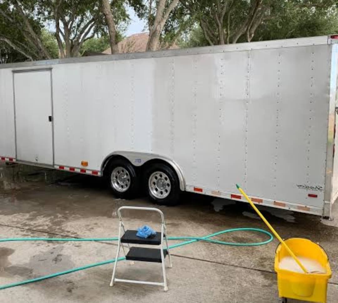 White enclosed trailer being cleaned on a driveway with a yellow bucket and step ladder.